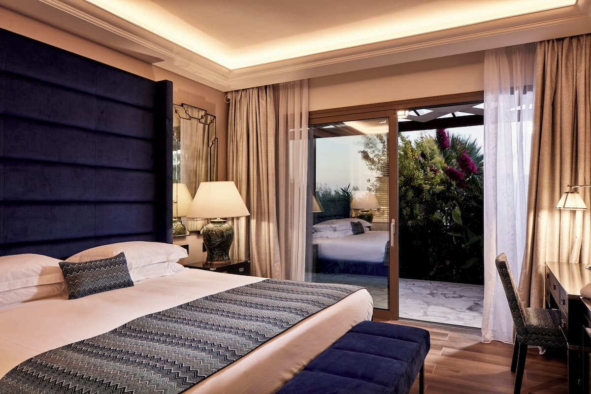 A hotel room with a large bed, a desk, a chair, a lamp, and a view through a glass door.