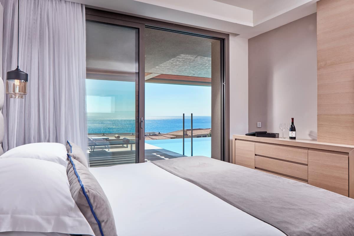 A bedroom with a bed, a nightstand, a bottle of wine, and a view of the ocean.