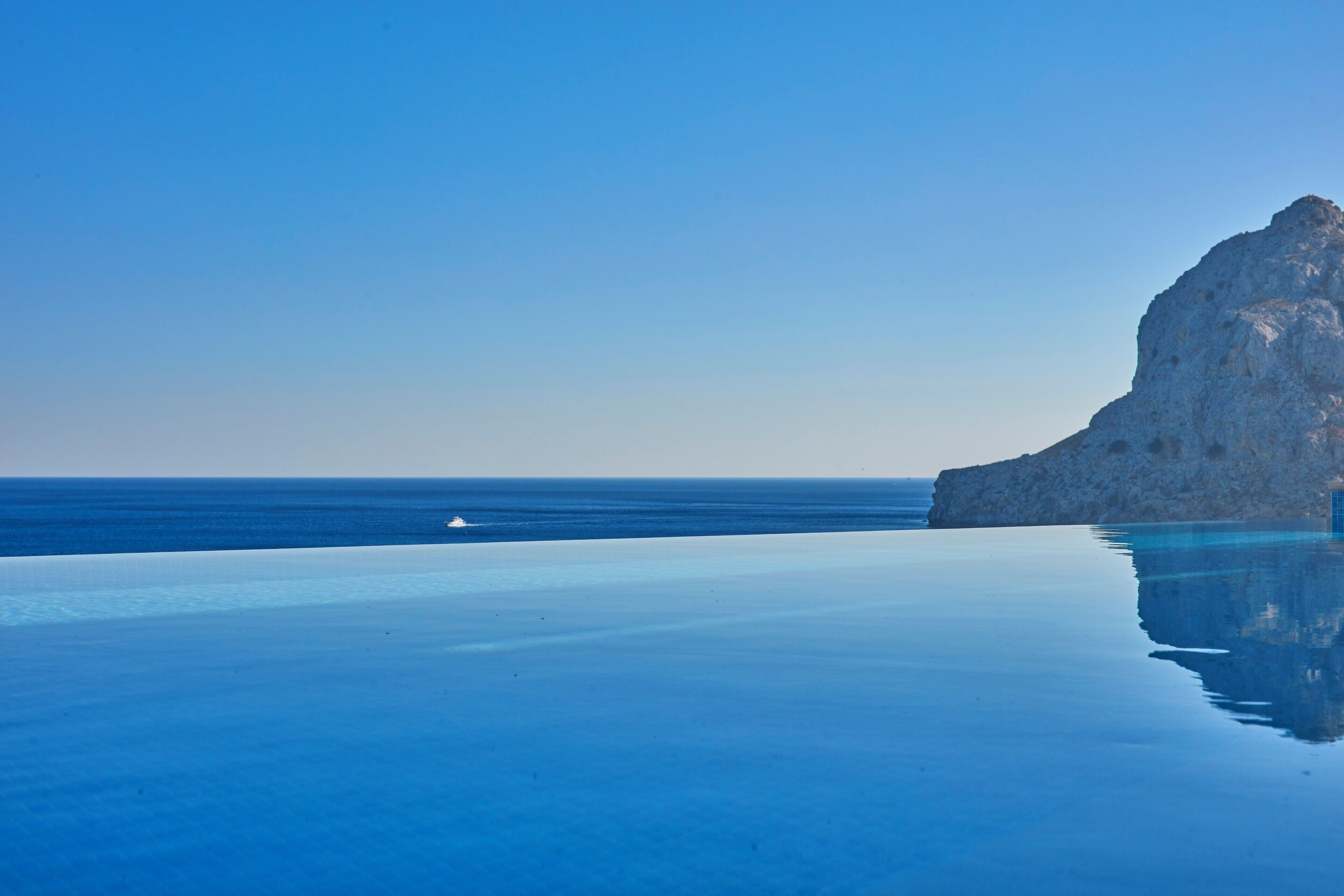 Piscina azul cristalina com um penhasco rochoso e o oceano ao fundo.