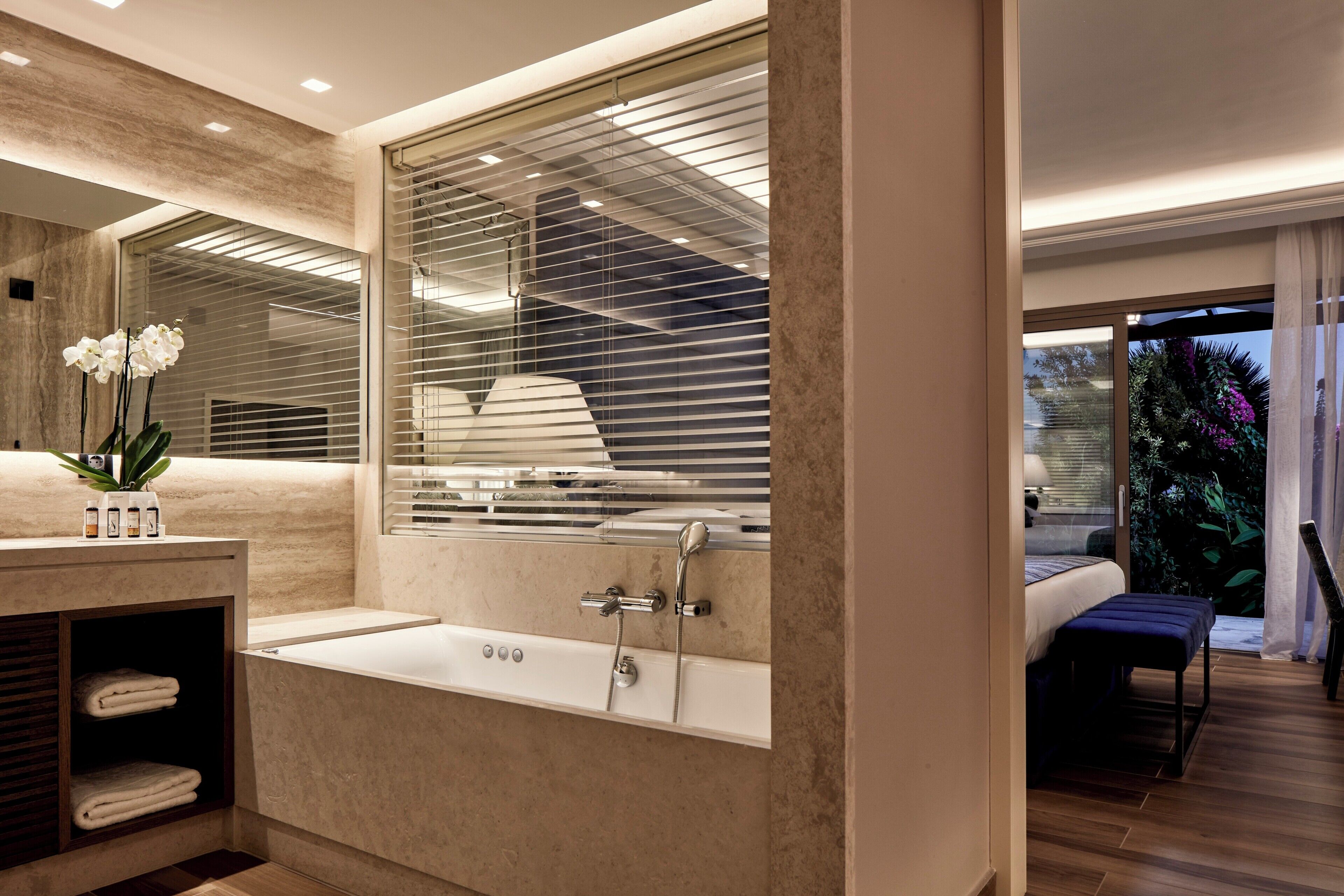 A modern bathroom with a bathtub, a large mirror, and a window with blinds.