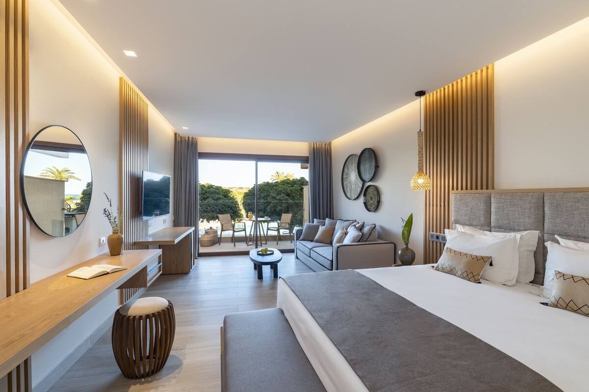 A modern hotel room with a large bed, a sofa, a dining area, and a view of the outdoors.