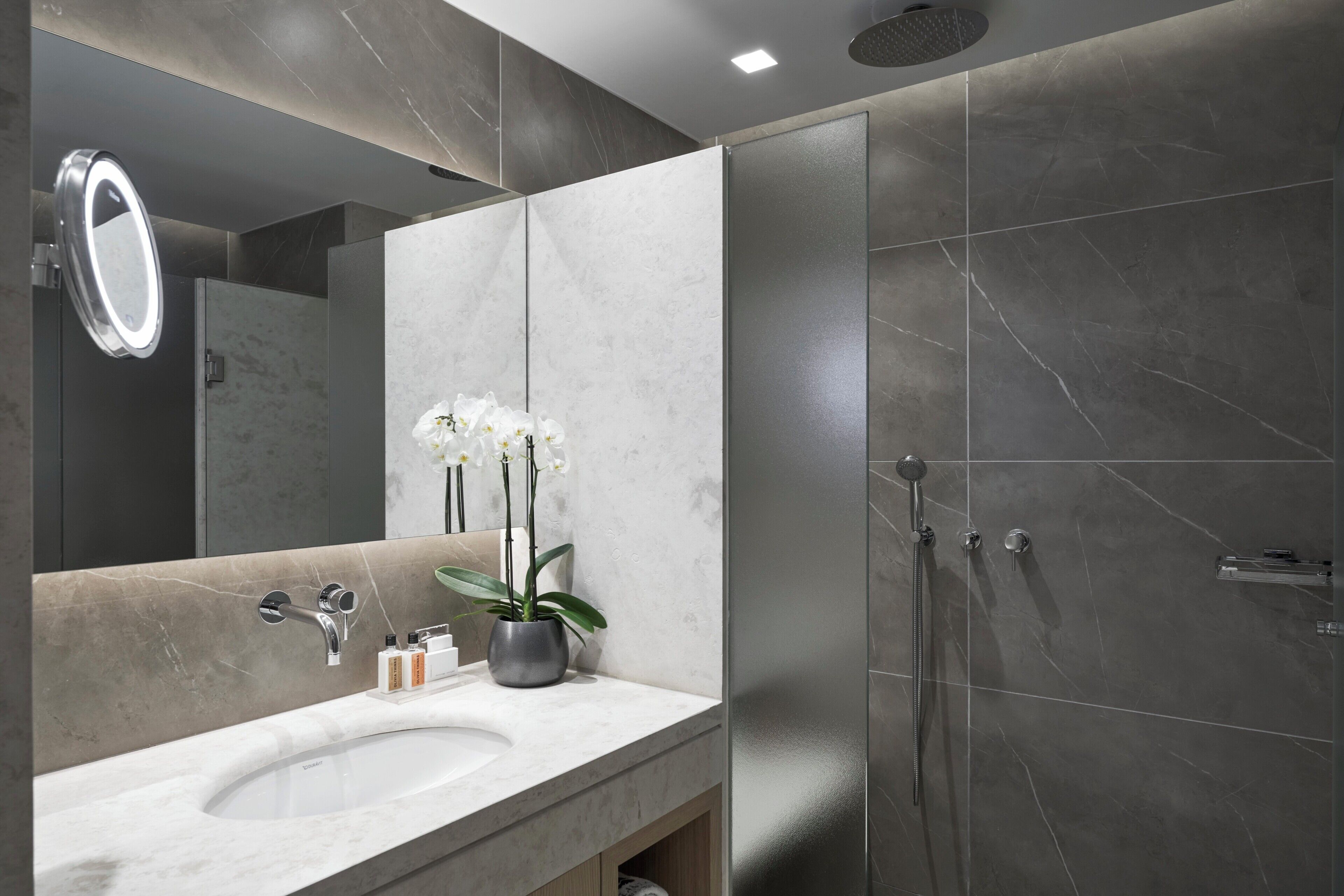 A modern bathroom with a marble sink, a mirror, a potted orchid, and a shower area.