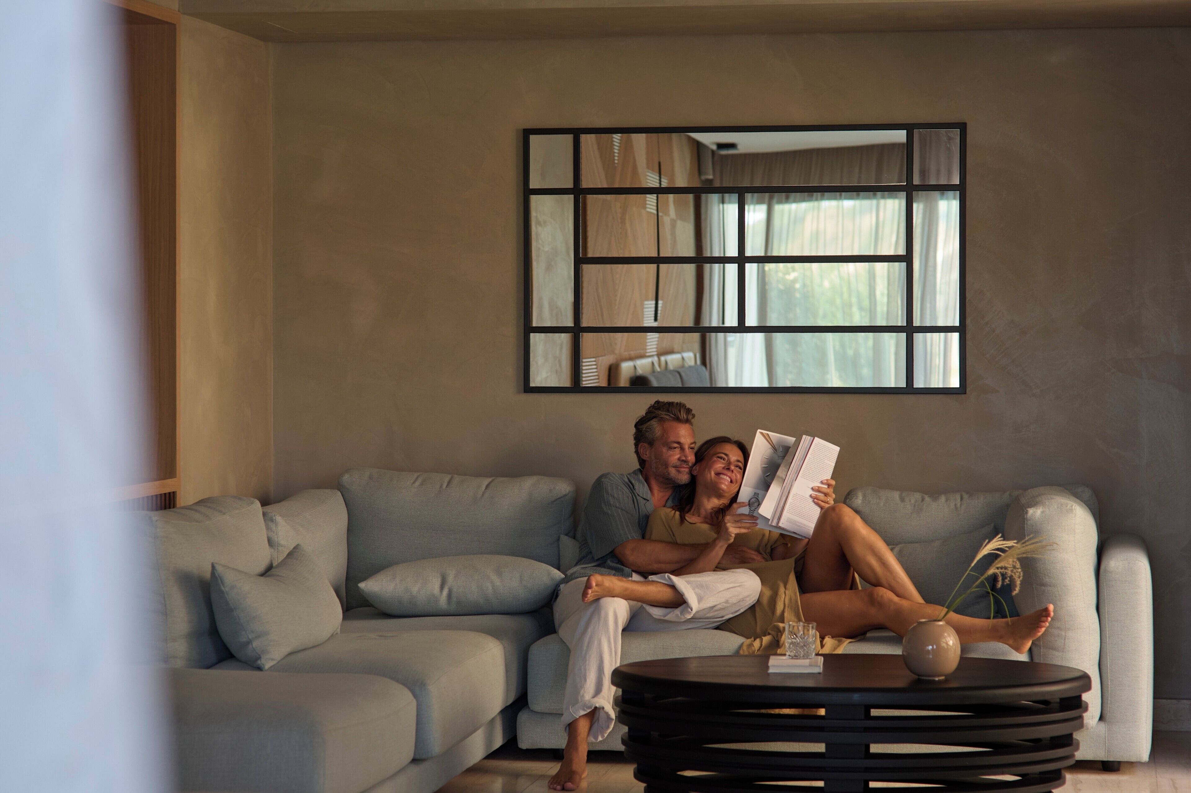 Una pareja en un sofá de tonos claros con una mesa de centro moderna, un espejo grande en la pared y una ventana con cortinas.