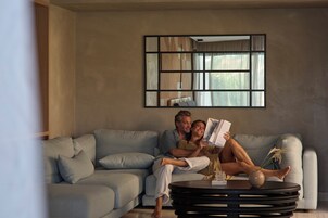 Una pareja en un sofá de tonos claros con una mesa de centro moderna, un espejo grande en la pared y una ventana con cortinas.