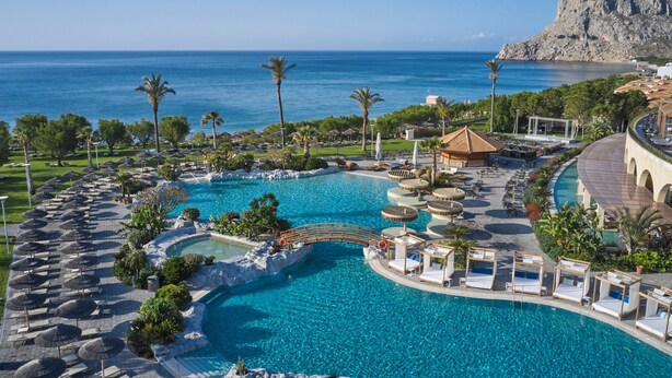 Une piscine d’un complexe hôtelier offrant une vue sur la mer, des palmiers et une montagne en arrière-plan.