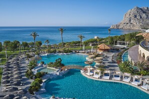 Zona de piscina en un resort con vista al mar, palmeras y una montaña al fondo.