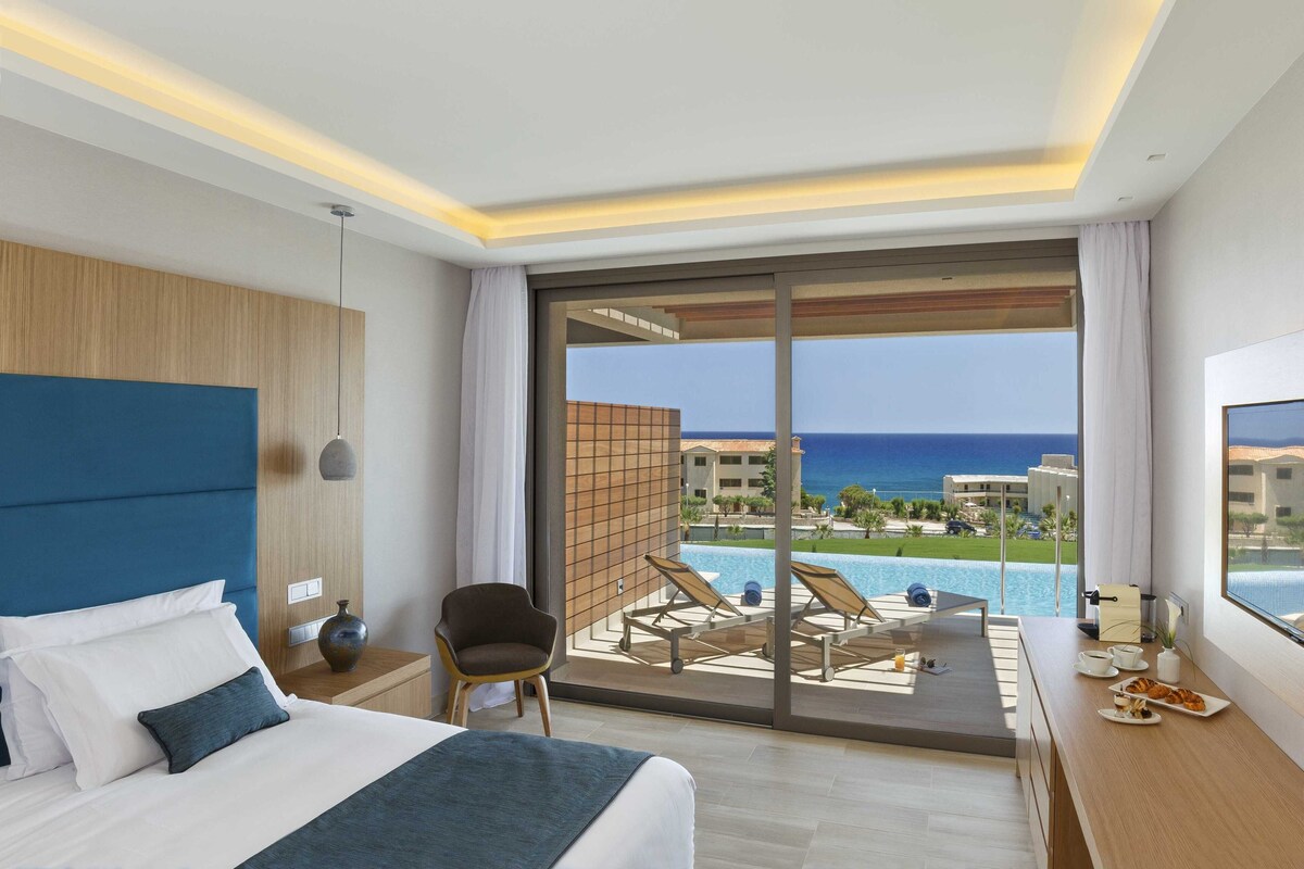 Une chambre d’hôtel comprenant un lit, une chaise, une table à manger avec des articles pour le petit-déjeuner et un balcon donnant sur la piscine.