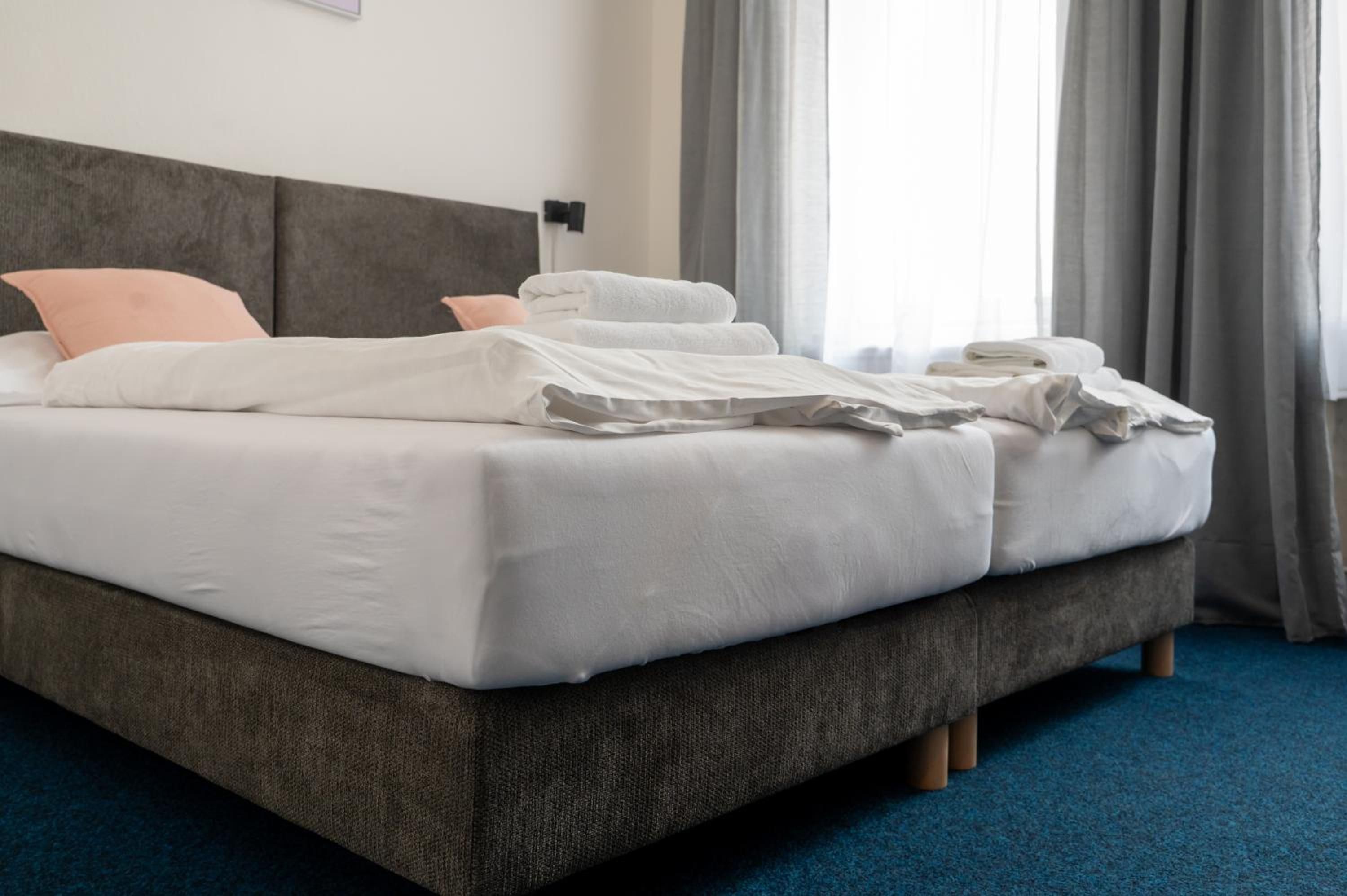 Un lit bien fait, avec du linge de lit blanc et des oreillers, une tête de lit rembourrée de couleur grise et un sol recouvert d’une moquette bleue.
