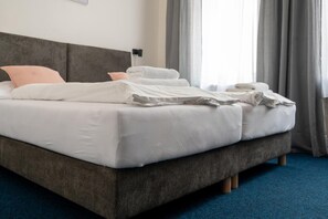 Een net opgemaakt bed met witte lakens en kussens, een grijs bekleed hoofdbord, en een blauw tapijt.