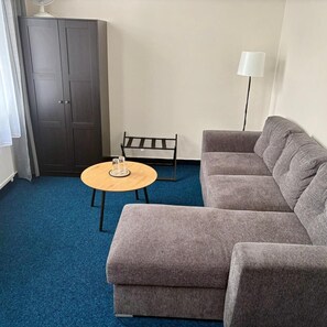 A room with a grey sofa, a round wooden table, a black chair, a dark wardrobe, and a floor lamp. - CB Royal (CESKE BUDEJOVICE)