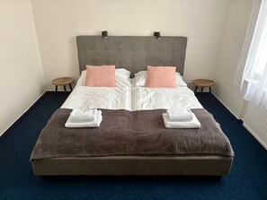 A double bed with a grey headboard, two pillows, two side tables, and two folded towels on a dark blue carpet.