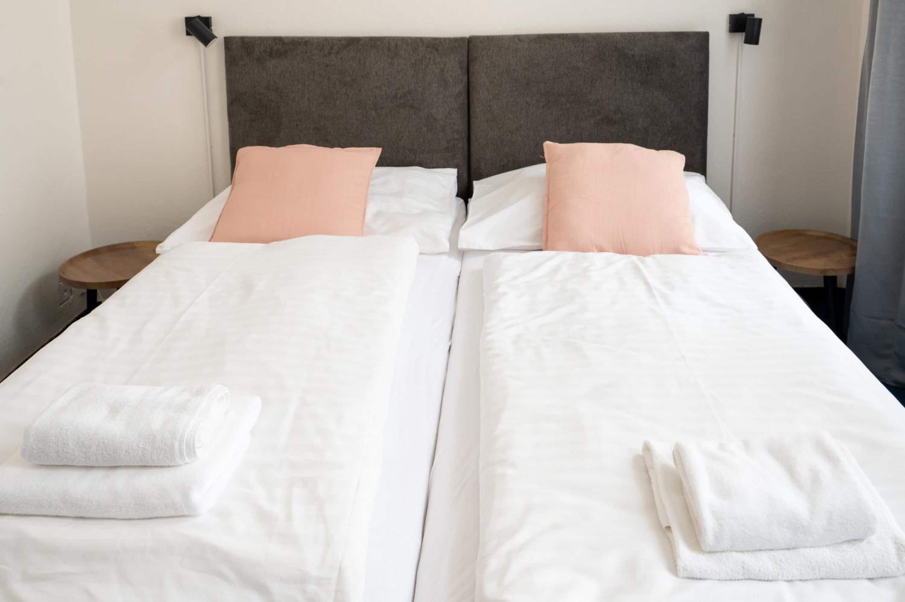 A double bed with white bedding and peach pillows, two bedside tables, and a grey headboard.