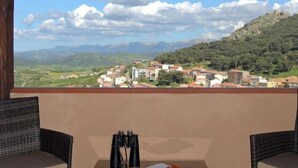A balcony with wicker furniture and binoculars, overlooking a town and mountains. - Su Lithu (Bitti)