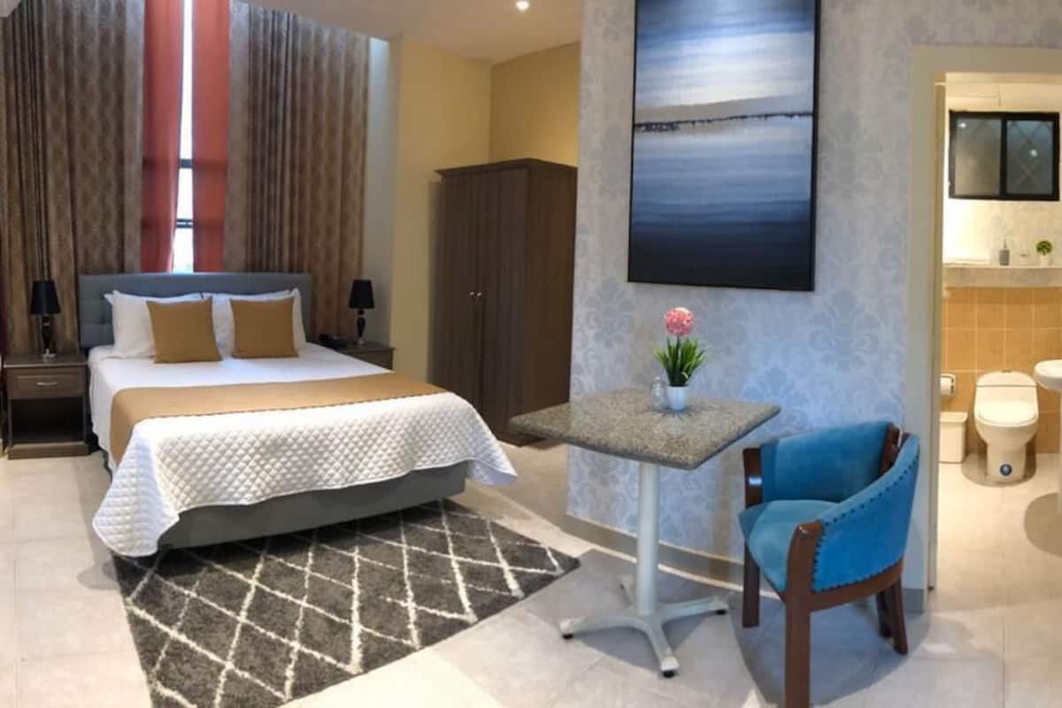 Habitación de hotel con cama, una mesita con un florero, una silla, armario, escritorio y baño visible a través de una puerta abierta.