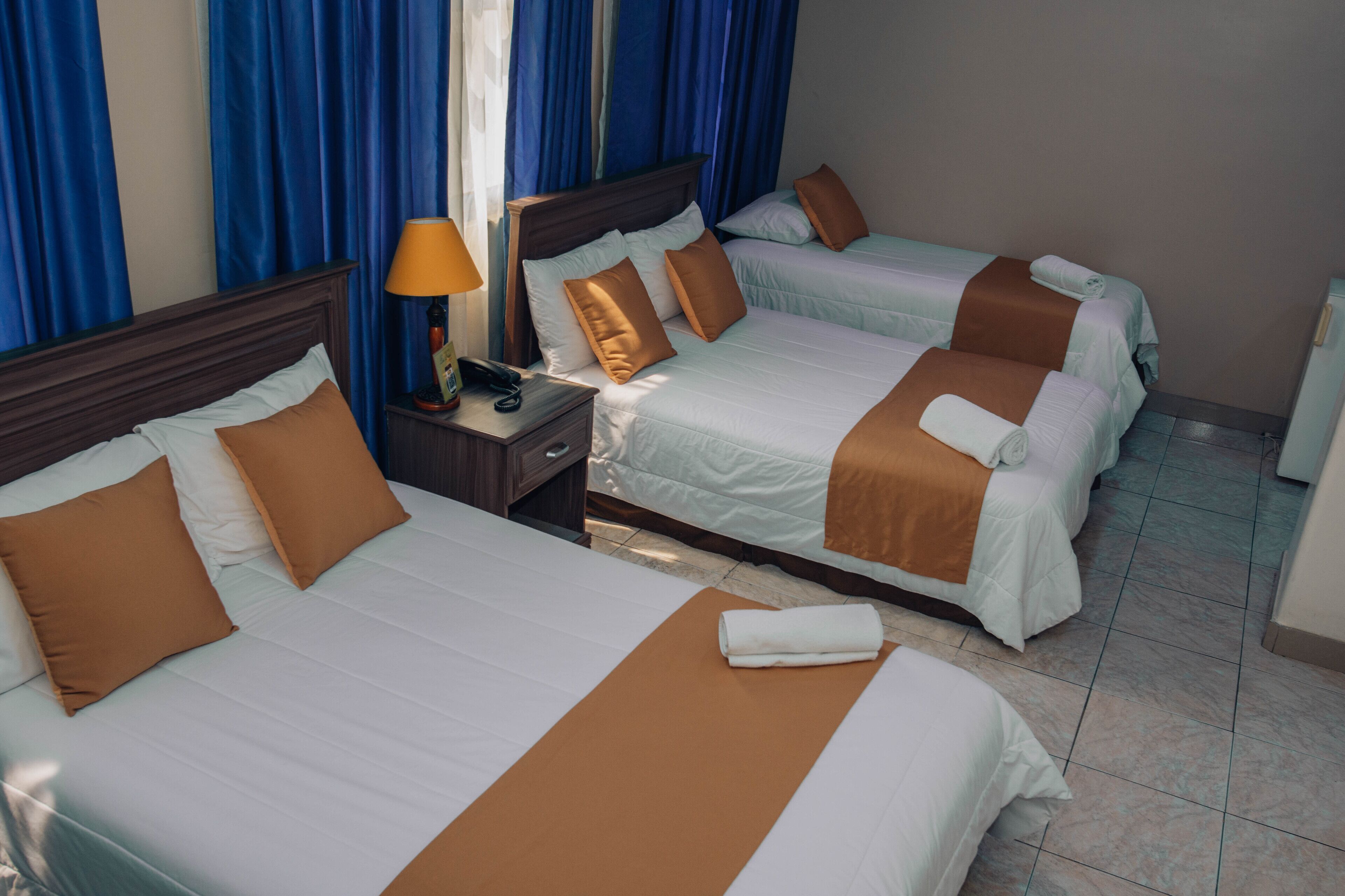A hotel room with three beds, each with white bedding and brown accents. There is a nightstand with a lamp and a telephone.