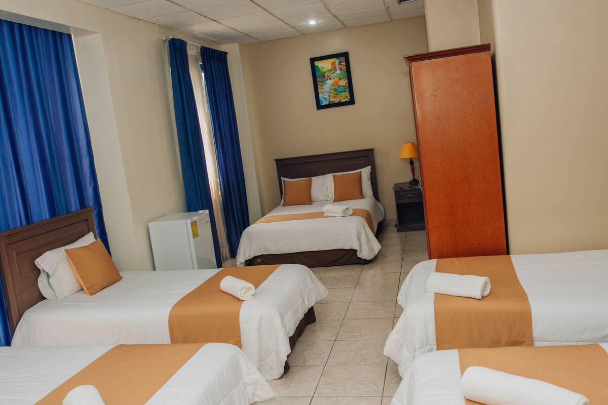 Habitación de hotel con cuatro camas, cada una con ropa de cama blanca y naranja, cabecero de madera y una mesita de noche pequeña con una lámpara.