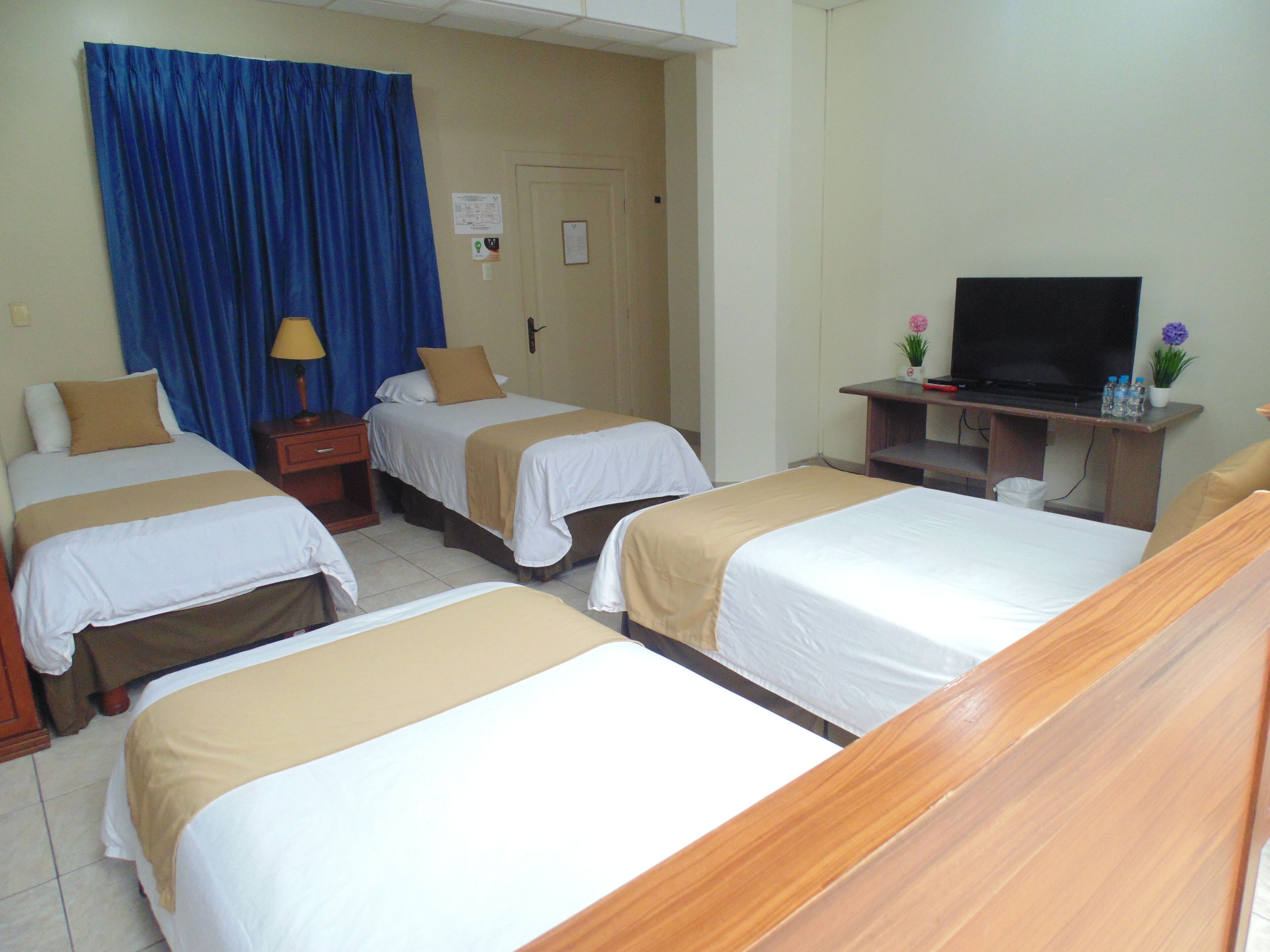 A hotel room with two single beds, a television on a wooden stand, a desk with a lamp, and a blue curtain.
