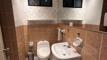 Un baño con inodoro, lavamanos y un cubo de basura. Hay dos ventanas con marcos negros.