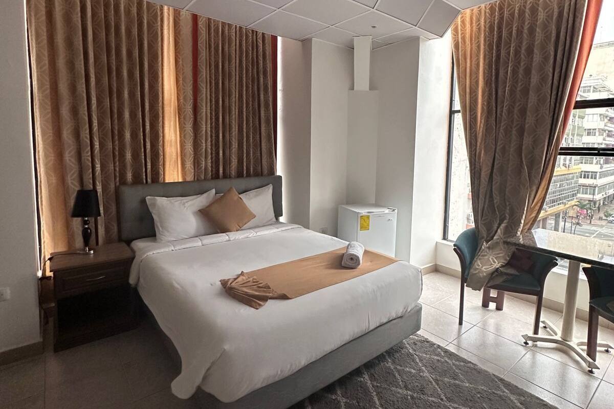 Una habitación de hotel con cama, mesita de noche, escritorio, silla y ventana con cortinas.