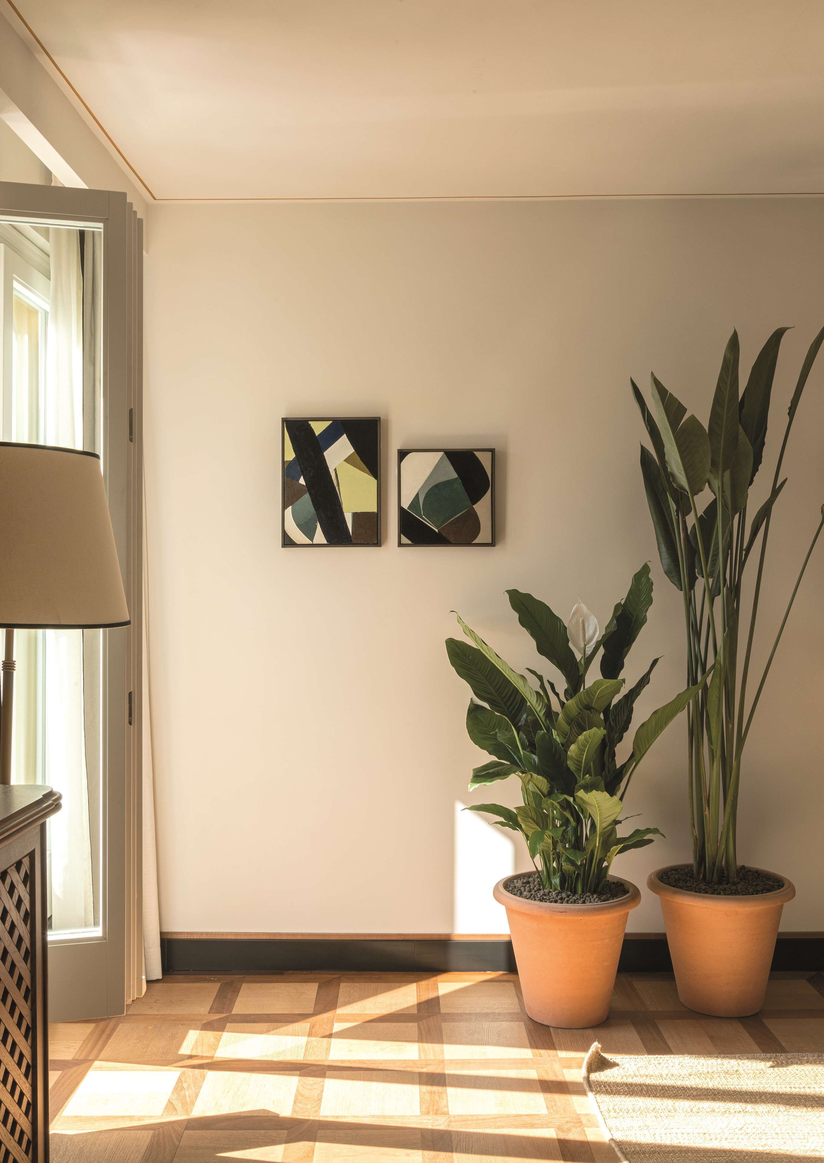 A room with a wooden floor, two potted plants, a lamp, and two abstract paintings on the wall.