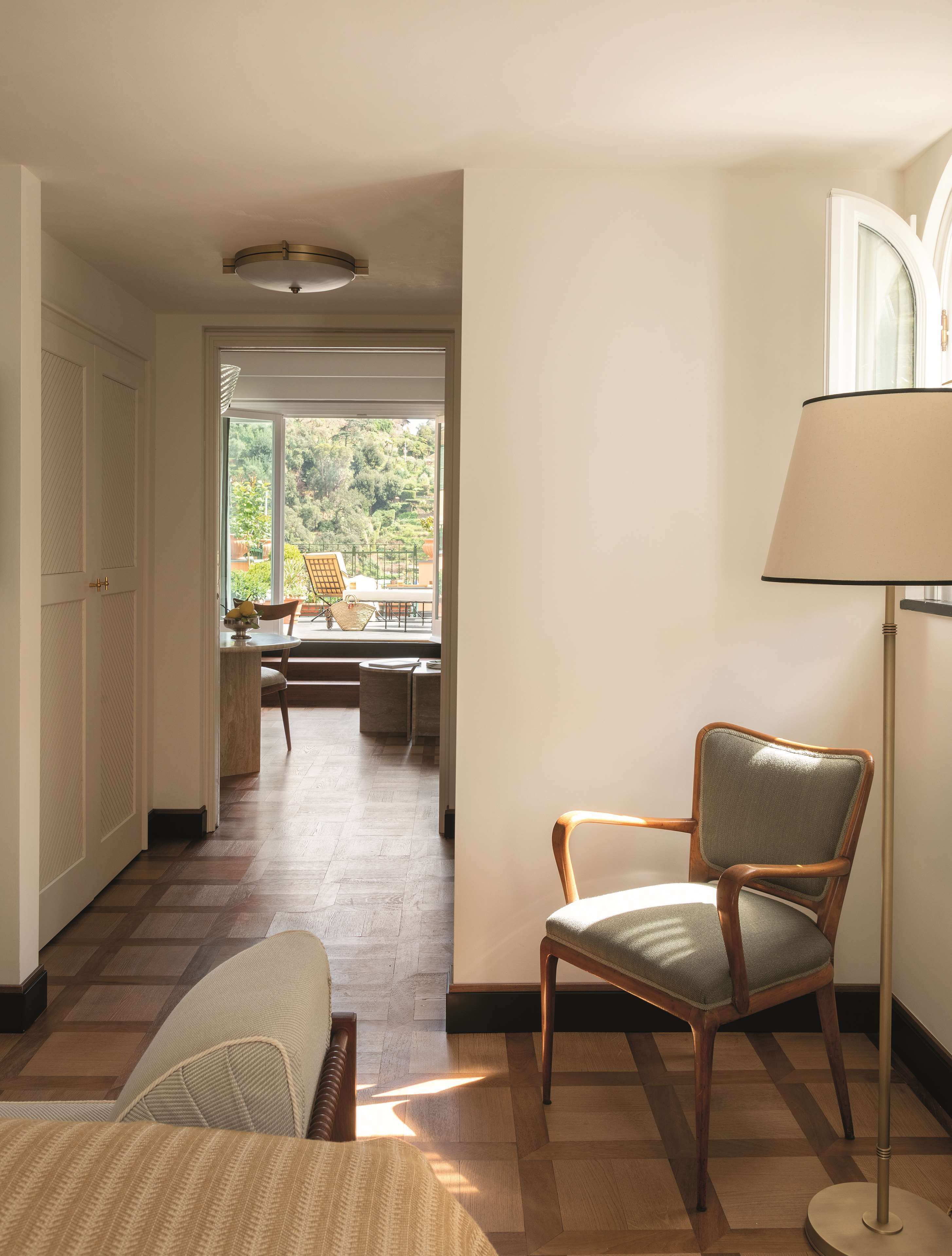 A room with a chair, a lamp, a window with a view of greenery, and a door leading to another room.
