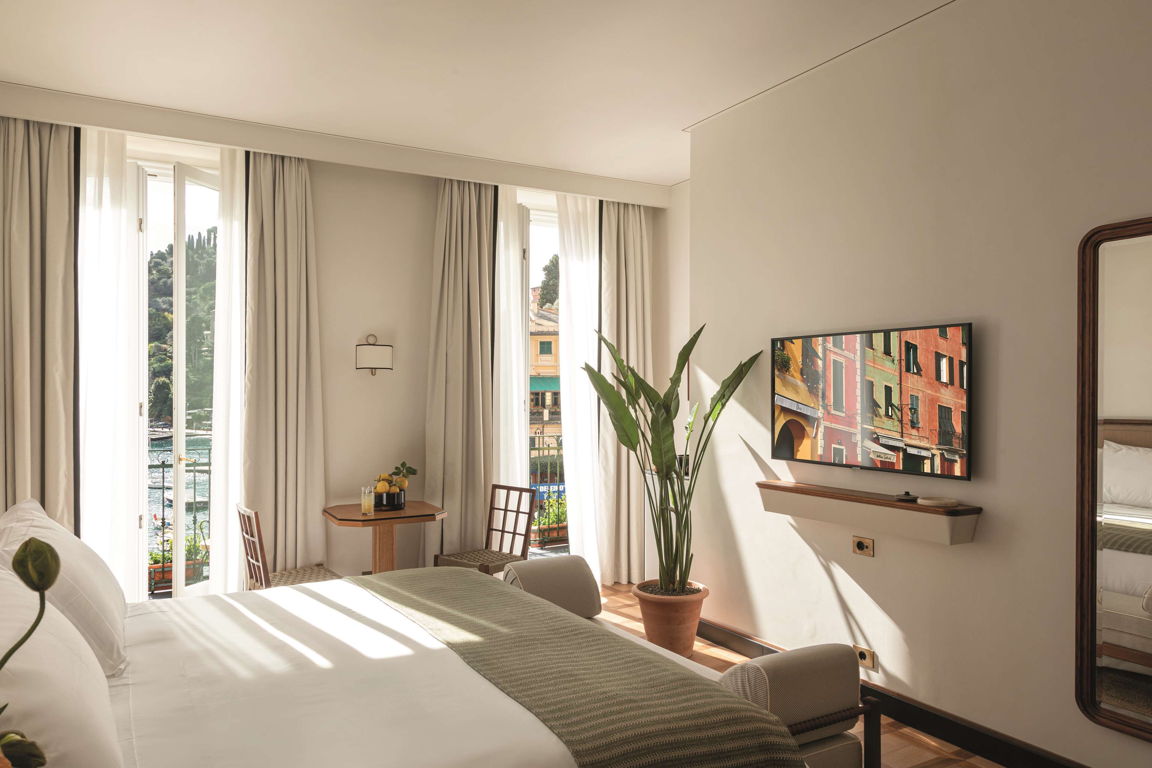 A hotel room with a bed, a chair, a small table, a potted plant, a TV, and a balcony with a view.