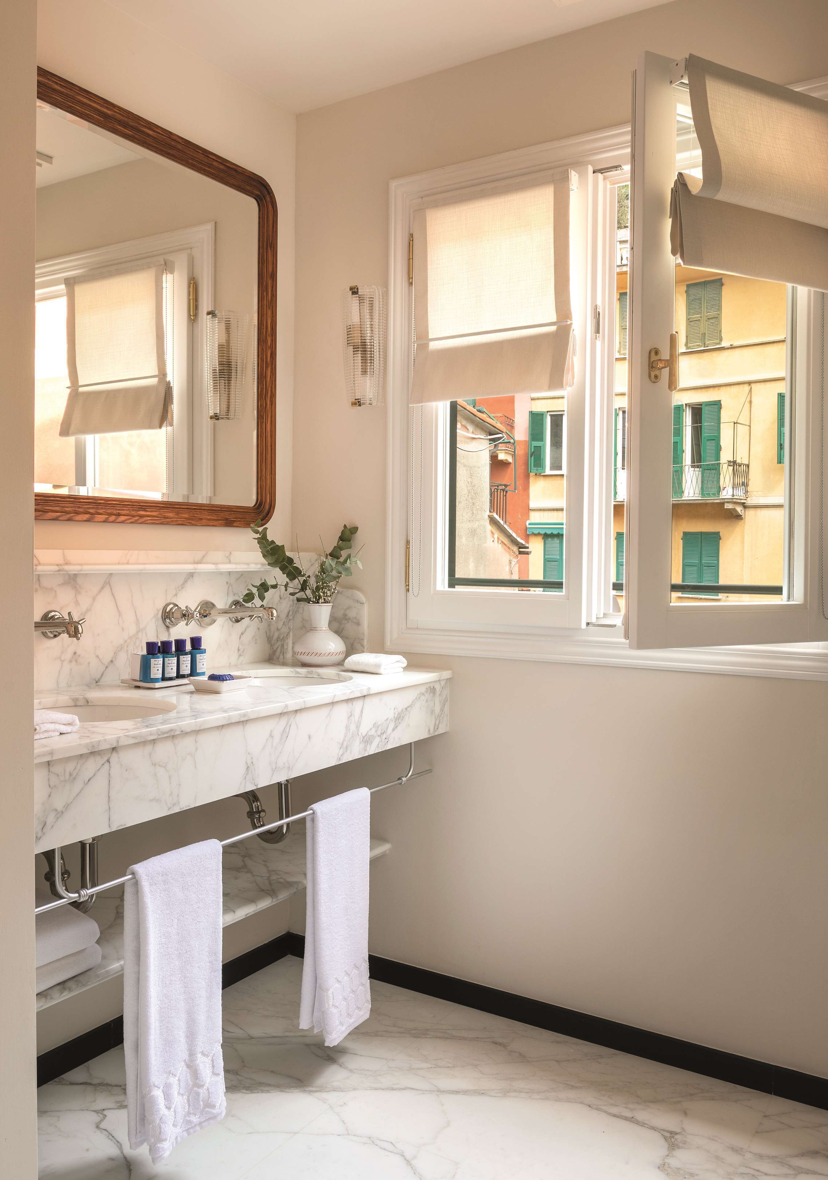 A bathroom with a marble sink, a mirror, a window with a view of buildings, and towels hanging on a rack.