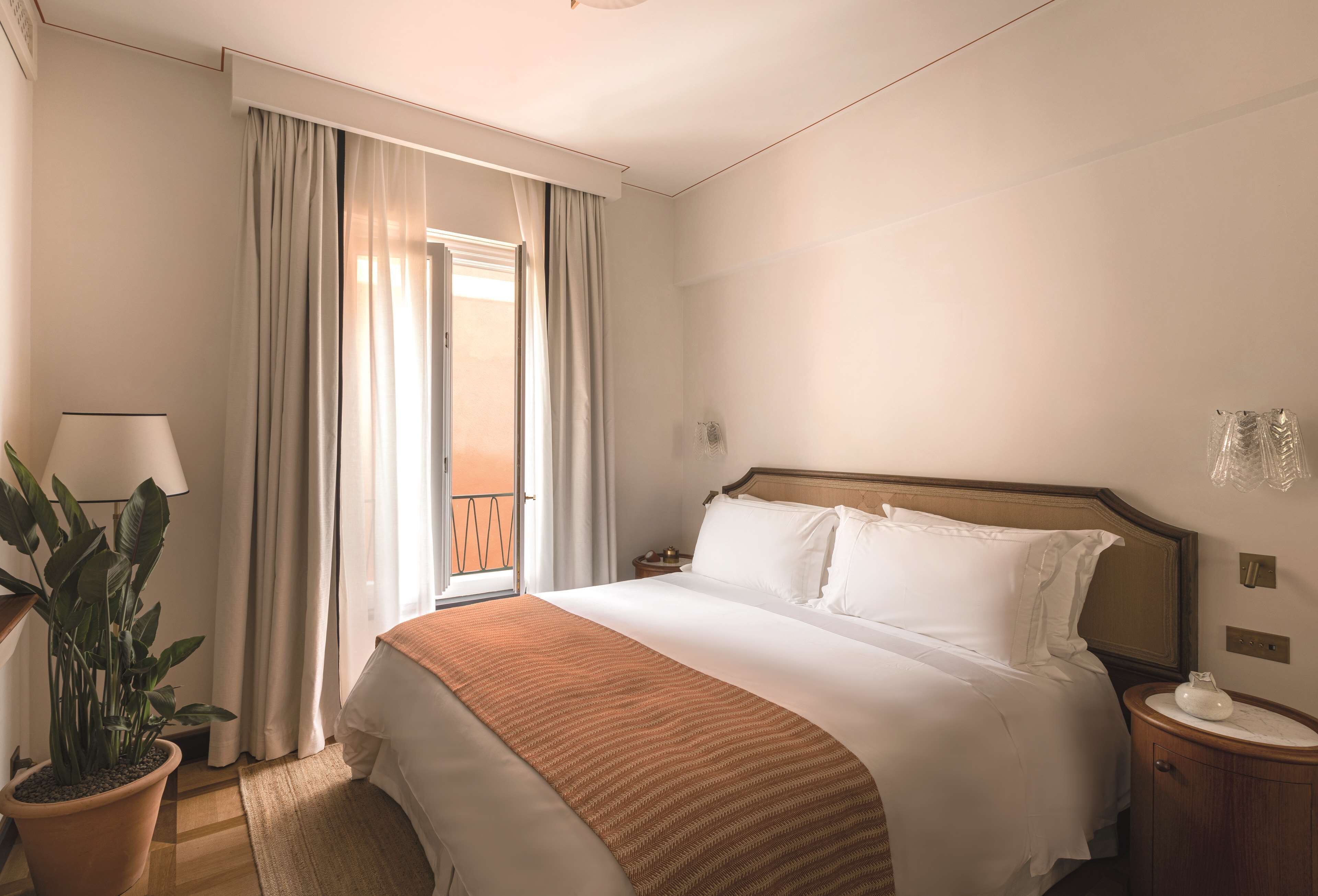 A hotel room with a large bed, a nightstand, a potted plant, and a view of the outdoors.
