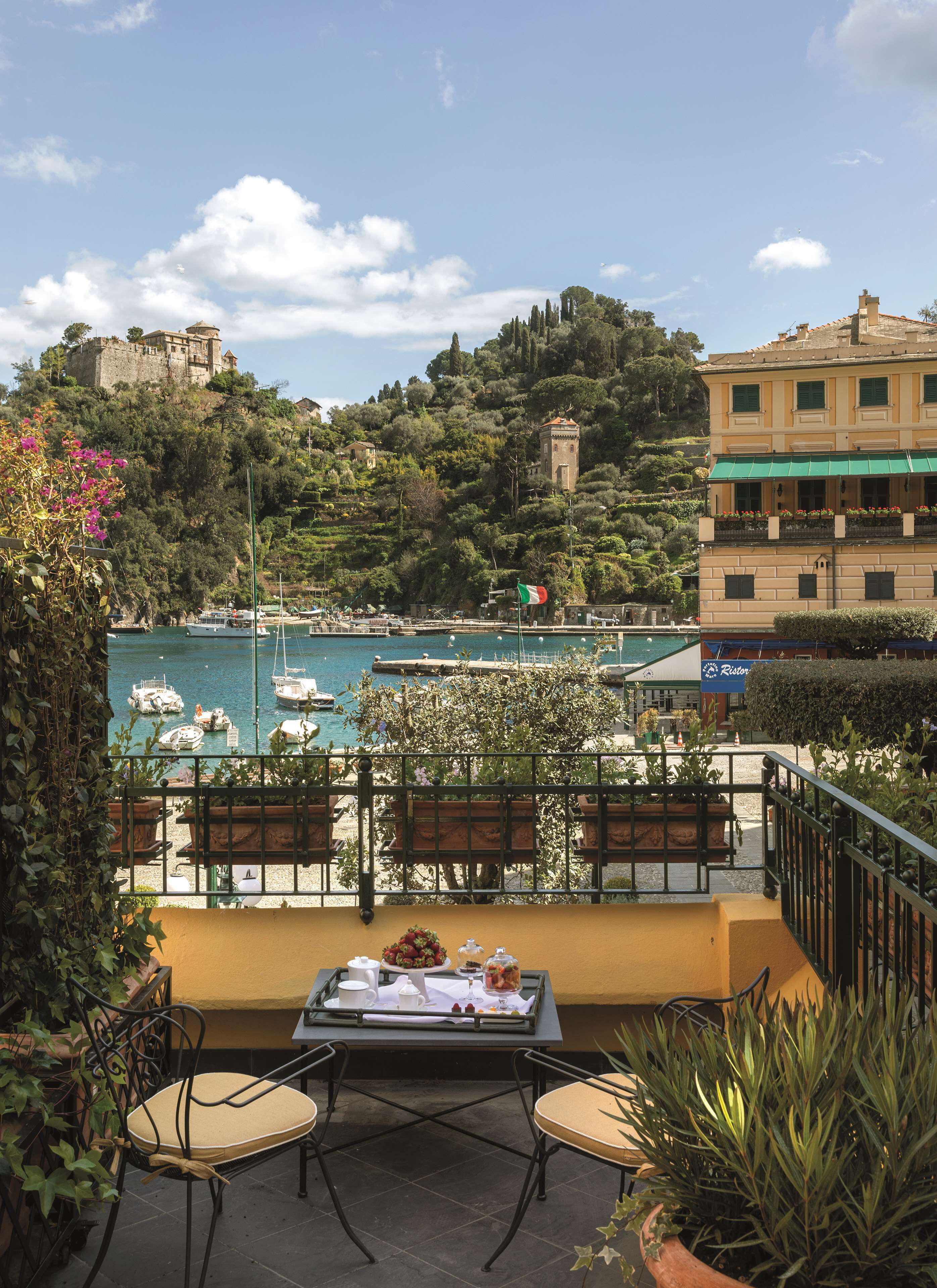 A terrace with a table set for two, overlooking a marina with boats and a hilltop building.