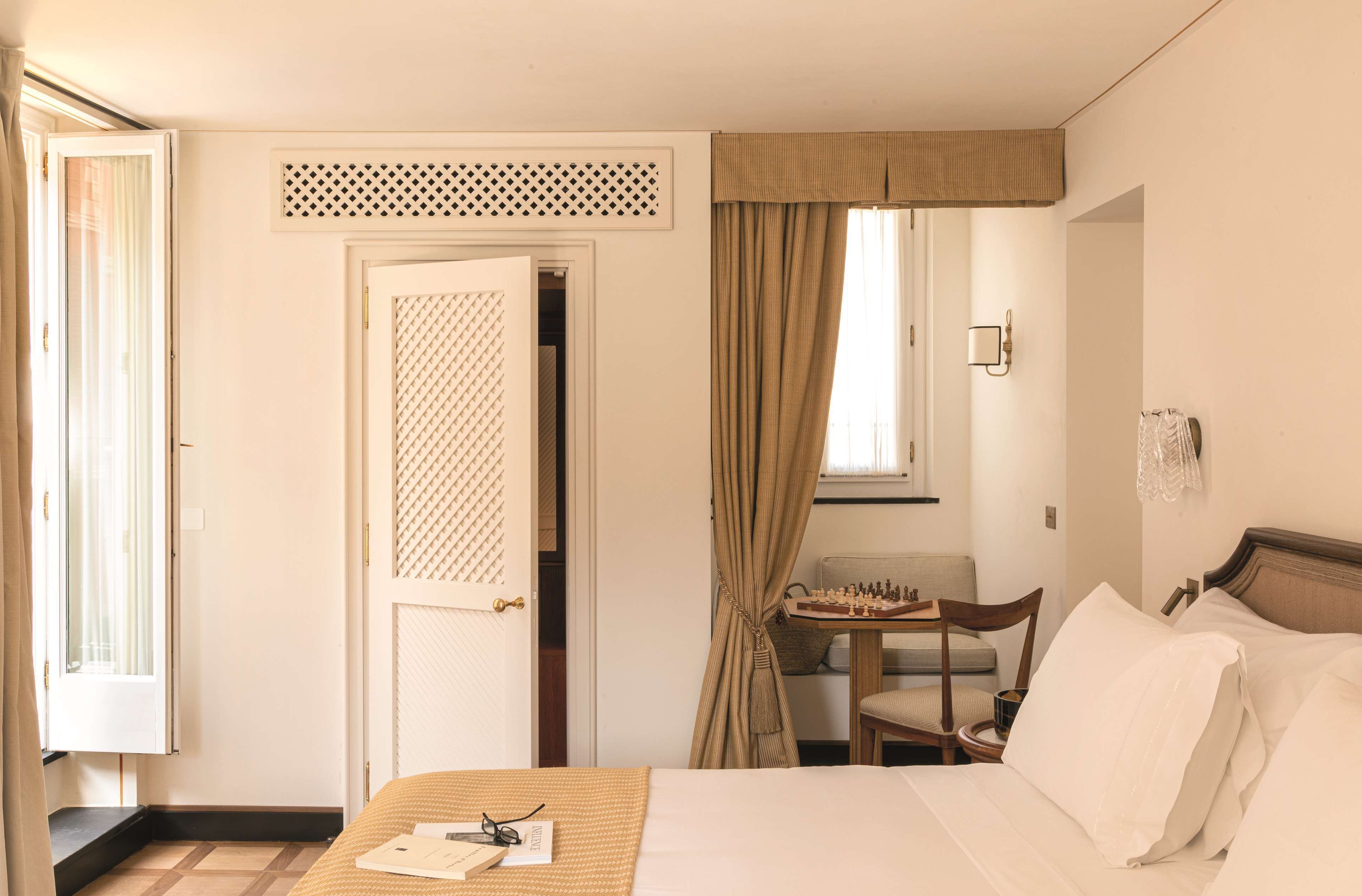 Quarto de hotel com uma cama, uma cadeira, uma mesinha com um tabuleiro de xadrez e uma janela com cortinas.