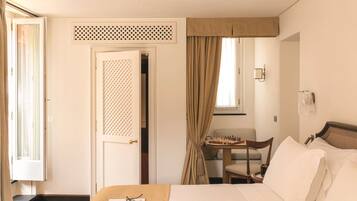 Quarto de hotel com uma cama, uma cadeira, uma mesinha com um tabuleiro de xadrez e uma janela com cortinas.