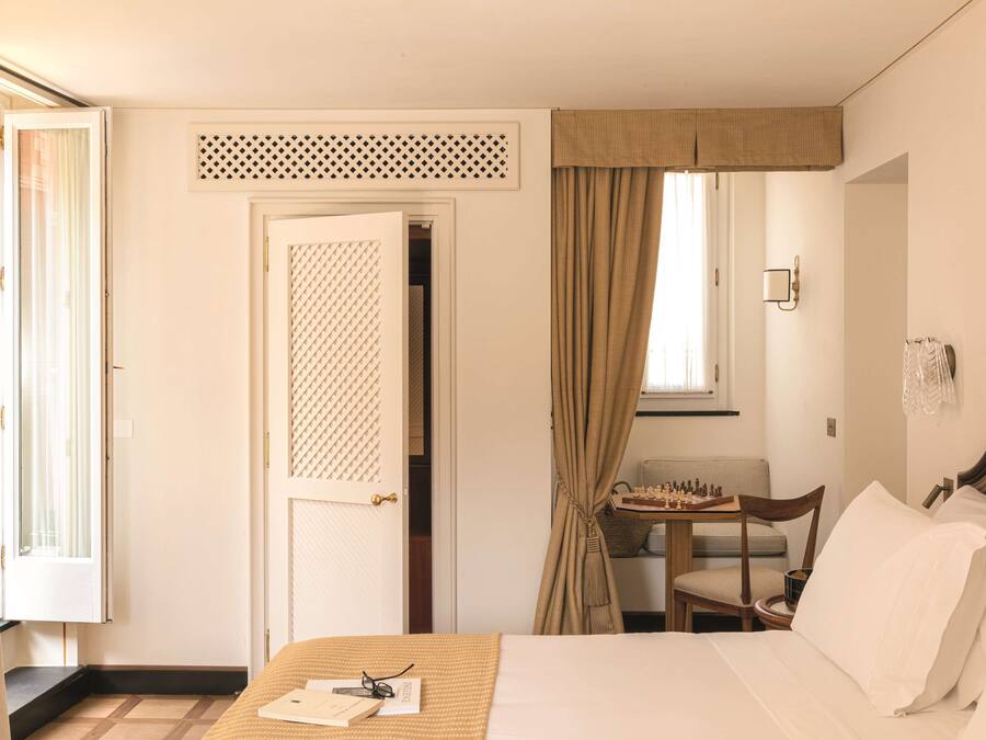 Quarto de hotel com uma cama, uma cadeira, uma mesinha com um tabuleiro de xadrez e uma janela com cortinas.
