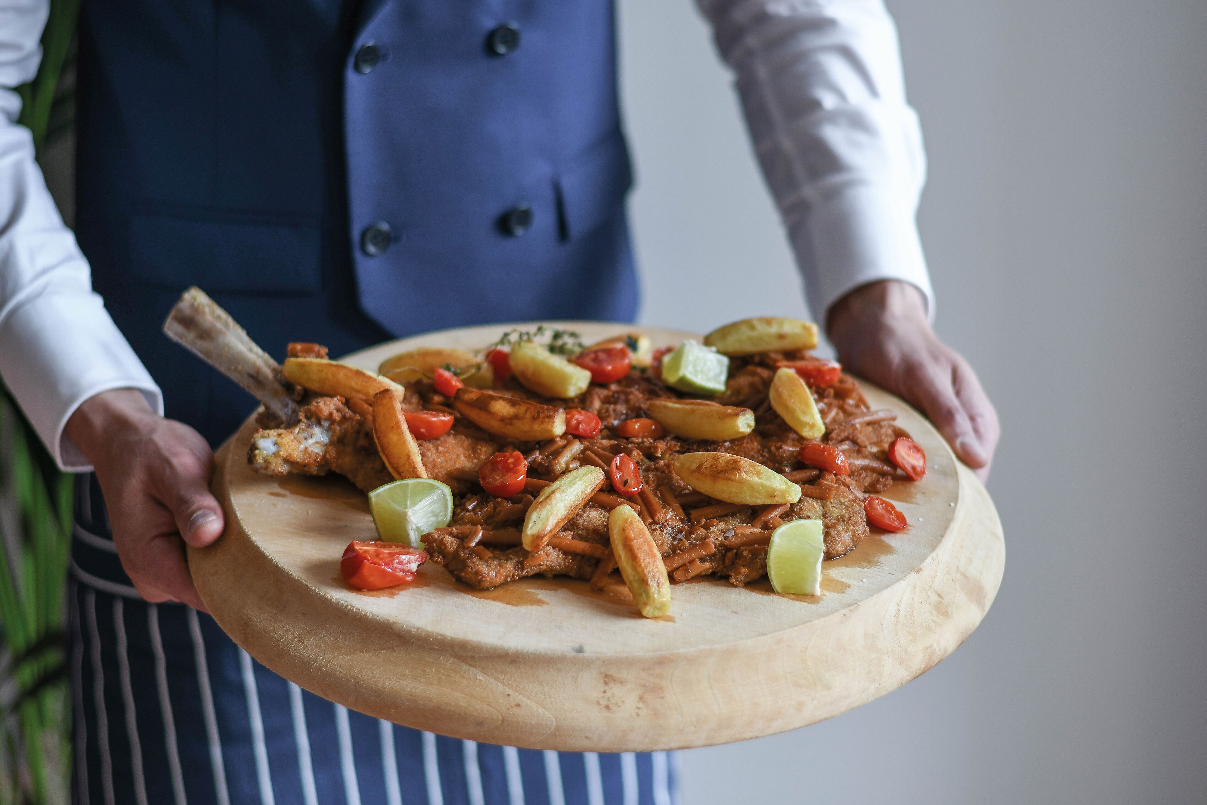 Una persona sosteniendo una tabla de madera con un plato de carne, papas y verduras.