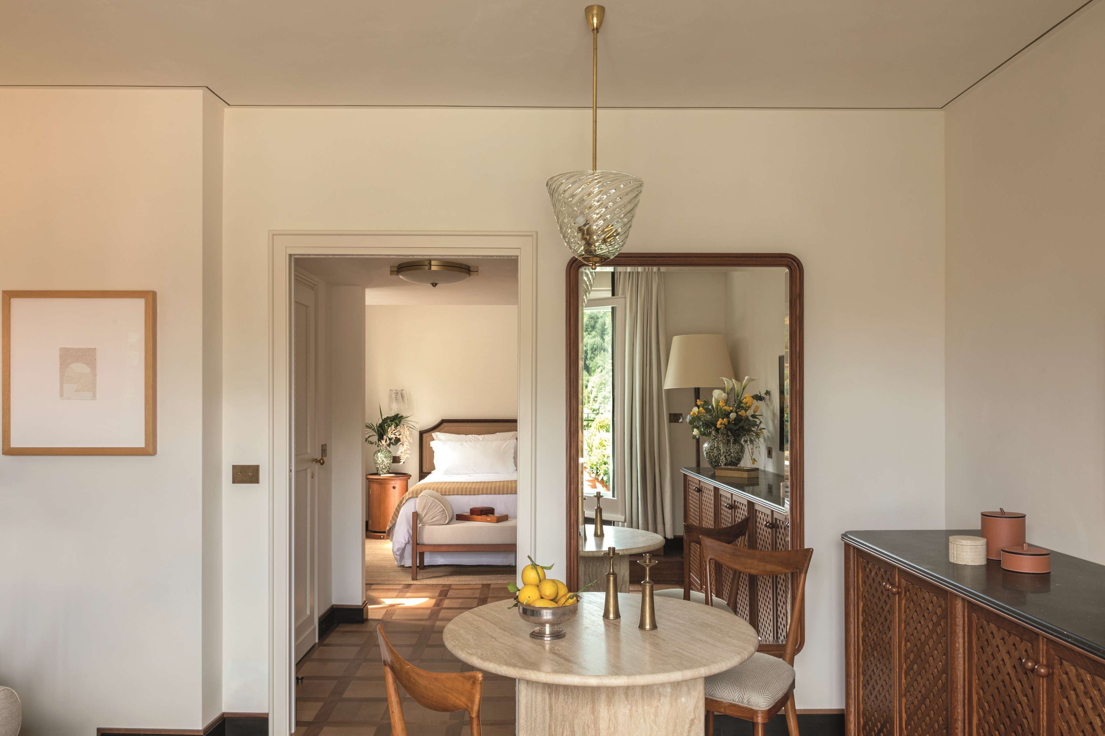 A hotel room with a round dining table, a bed with a wooden headboard, a mirror, and a hallway leading to another room.