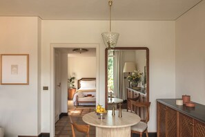 A hotel room with a round dining table, a bed with a wooden headboard, a mirror, and a hallway leading to another room.