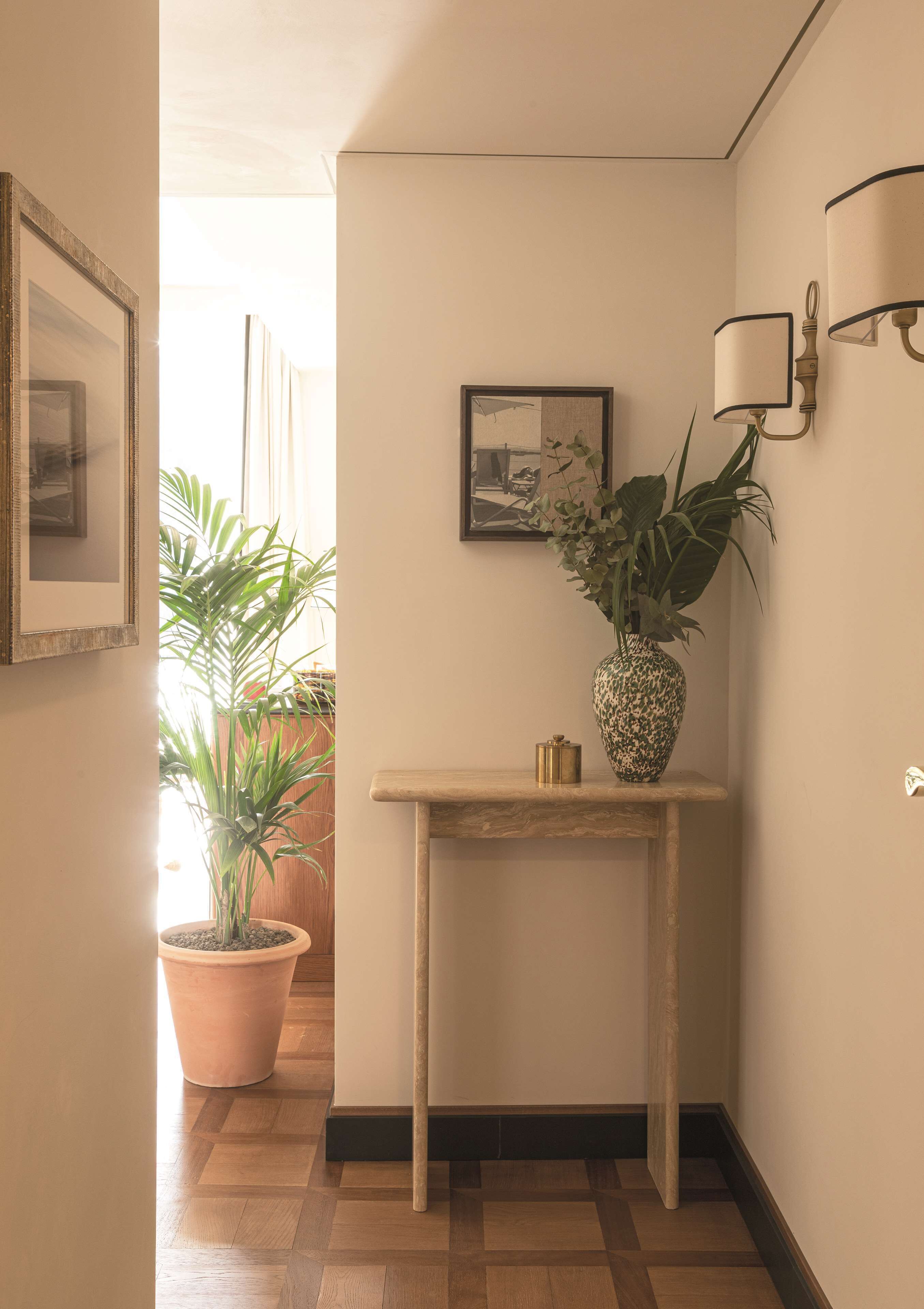 Un pasillo con suelo de madera, una mesita con planta en maceta, un cuadro enmarcado y una lámpara fijada a la pared.