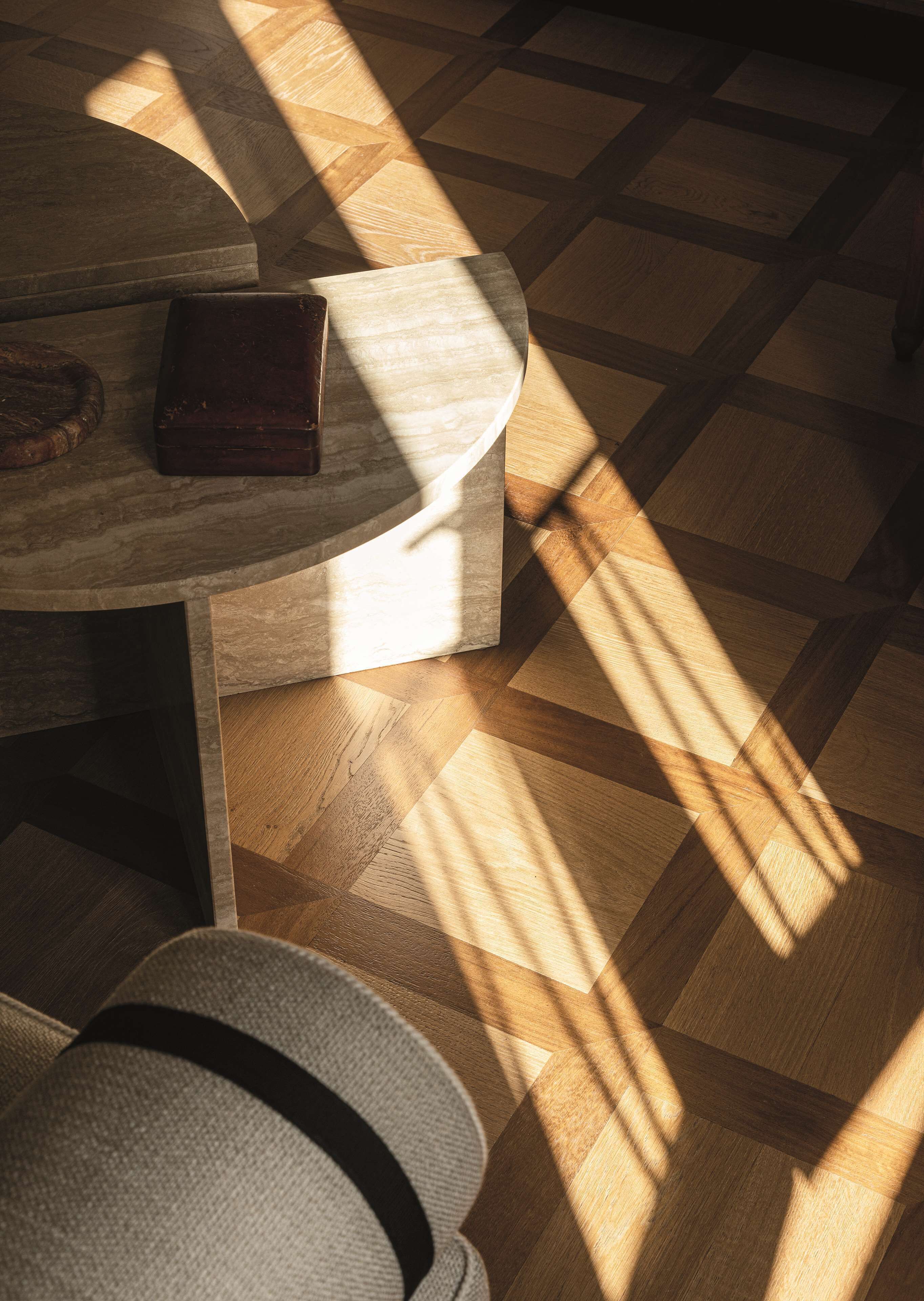 Ein Holzboden mit geometrischem Muster, ein runder Tisch mit einem dunklen Gegenstand darauf und ein Teil eines Stuhls.