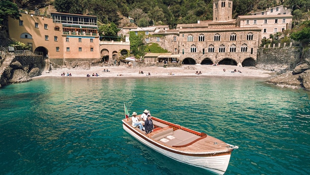 Un bote en un mar turquesa con un edificio histórico y un litoral rocoso al fondo.