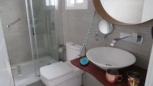 Un baño moderno con cabina de ducha de vidrio, un inodoro blanco, un lavamanos grande con encimera de madera y un espejo redondo.