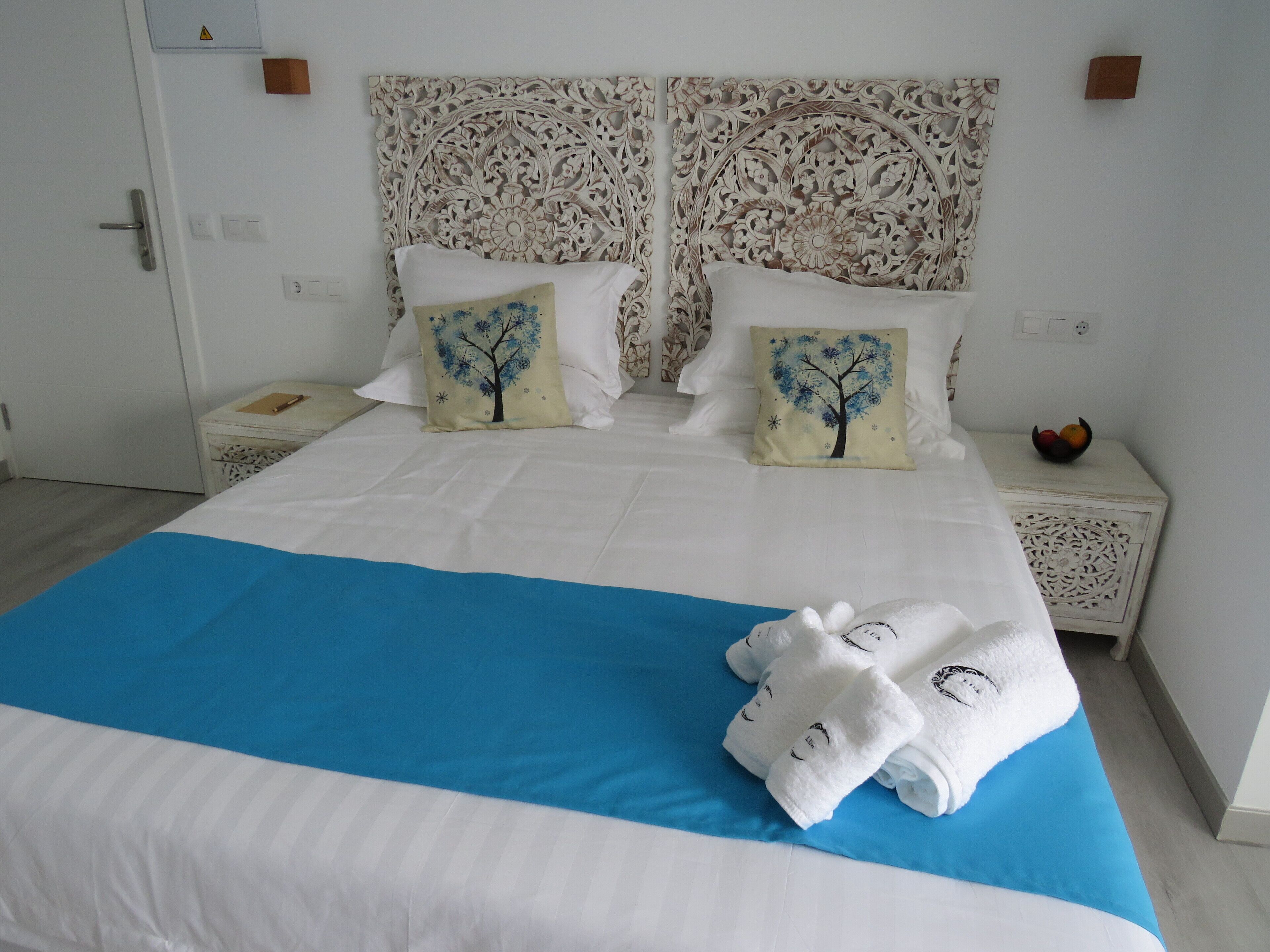 A neatly made bed with white and blue bedding, two pillows with tree designs, a white nightstand with a fruit bowl, and a decorative headboard.