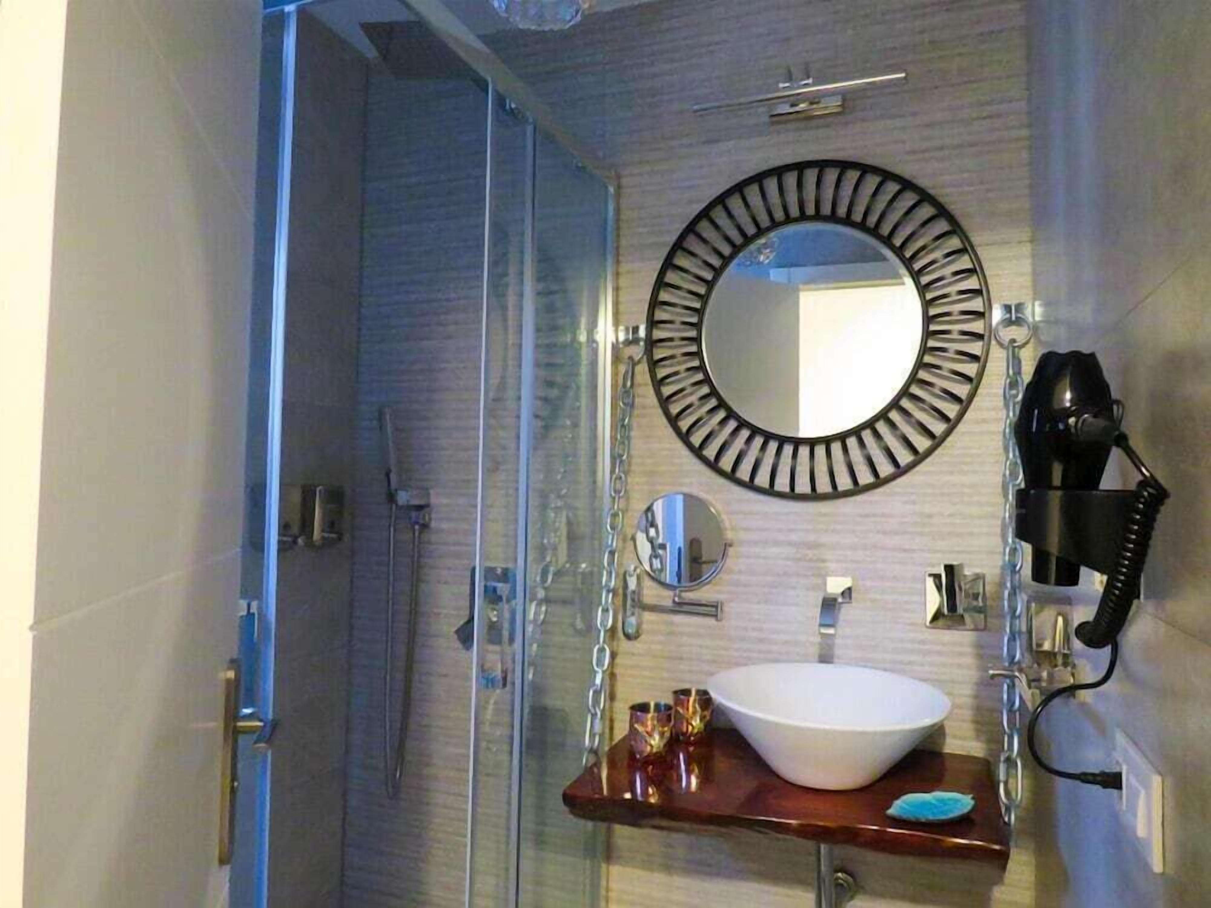 A bathroom with a glass shower enclosure, a round mirror with a black frame, a white sink with a wooden countertop, and a hairdryer on the wall.