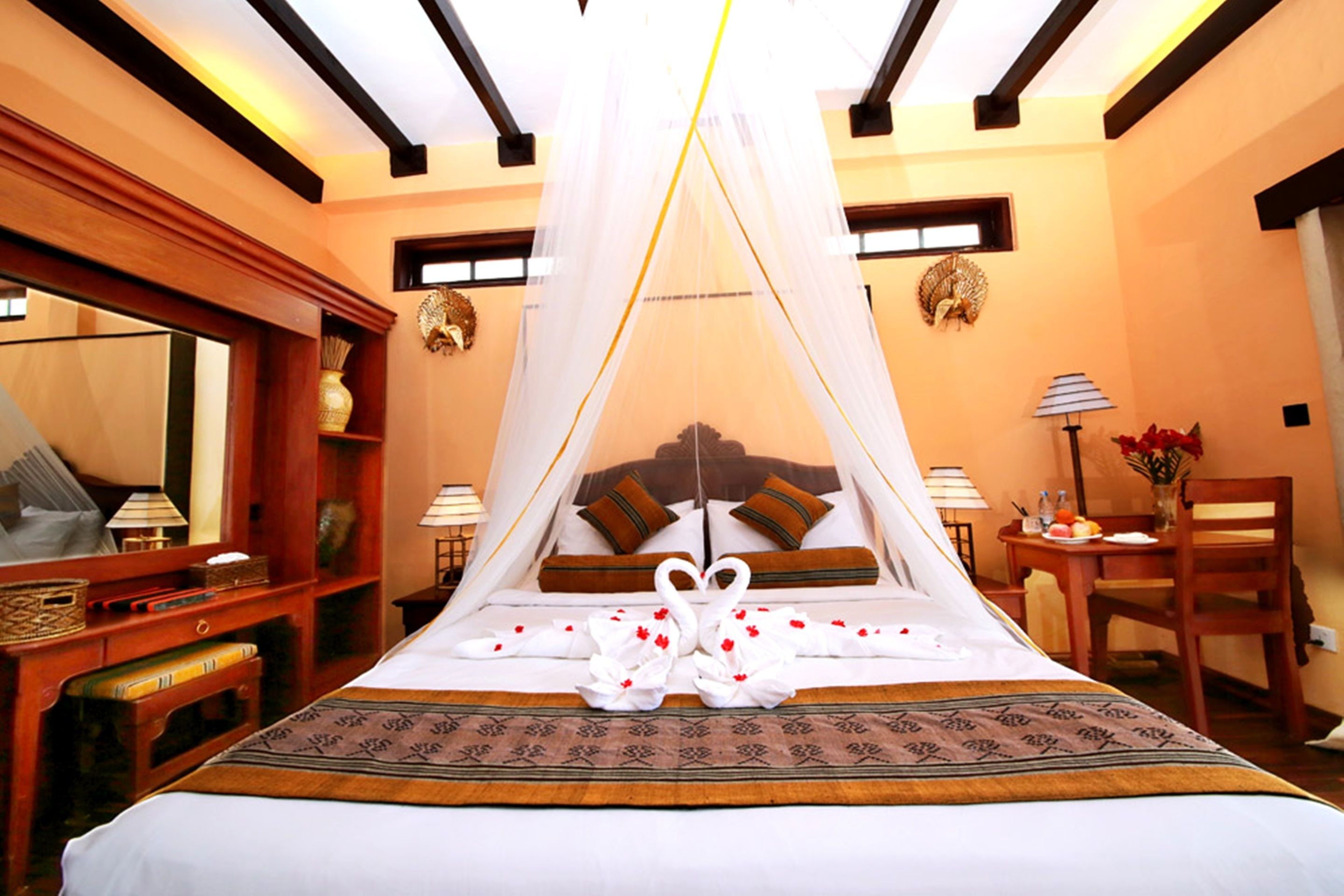 A hotel room with a large bed, a canopy bed, a wooden desk, a chair, a table with a vase of flowers, and a mirror.
