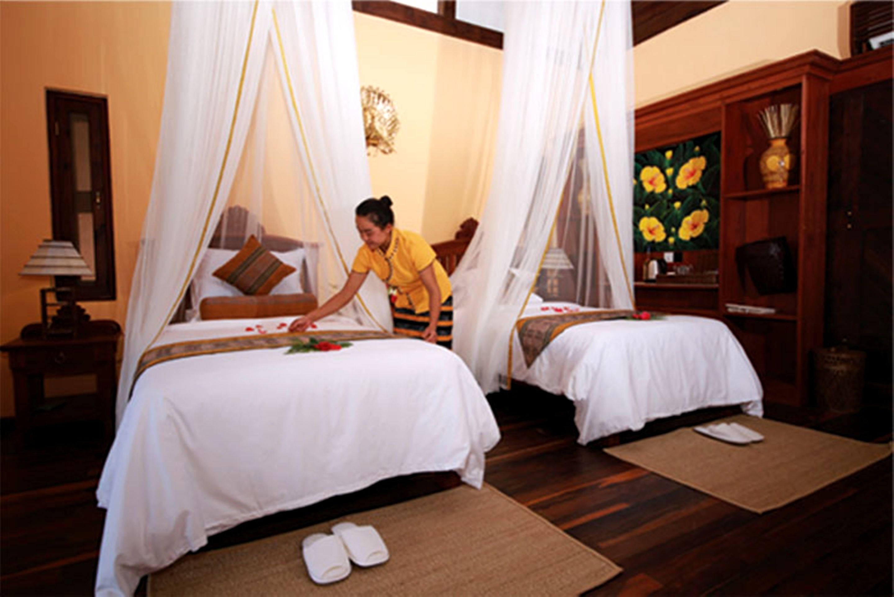 A room with two beds, each with a canopy, a wooden cabinet with a floral painting, and slippers on the floor.