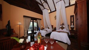A hotel room with two beds, a dining table with fruit and wine glasses, a TV, and a window with curtains.