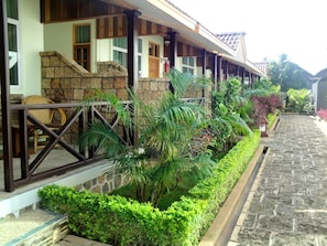 Un hotel con fachada de piedra, un espacio exterior cubierto con barandilla de madera y abundante vegetación.