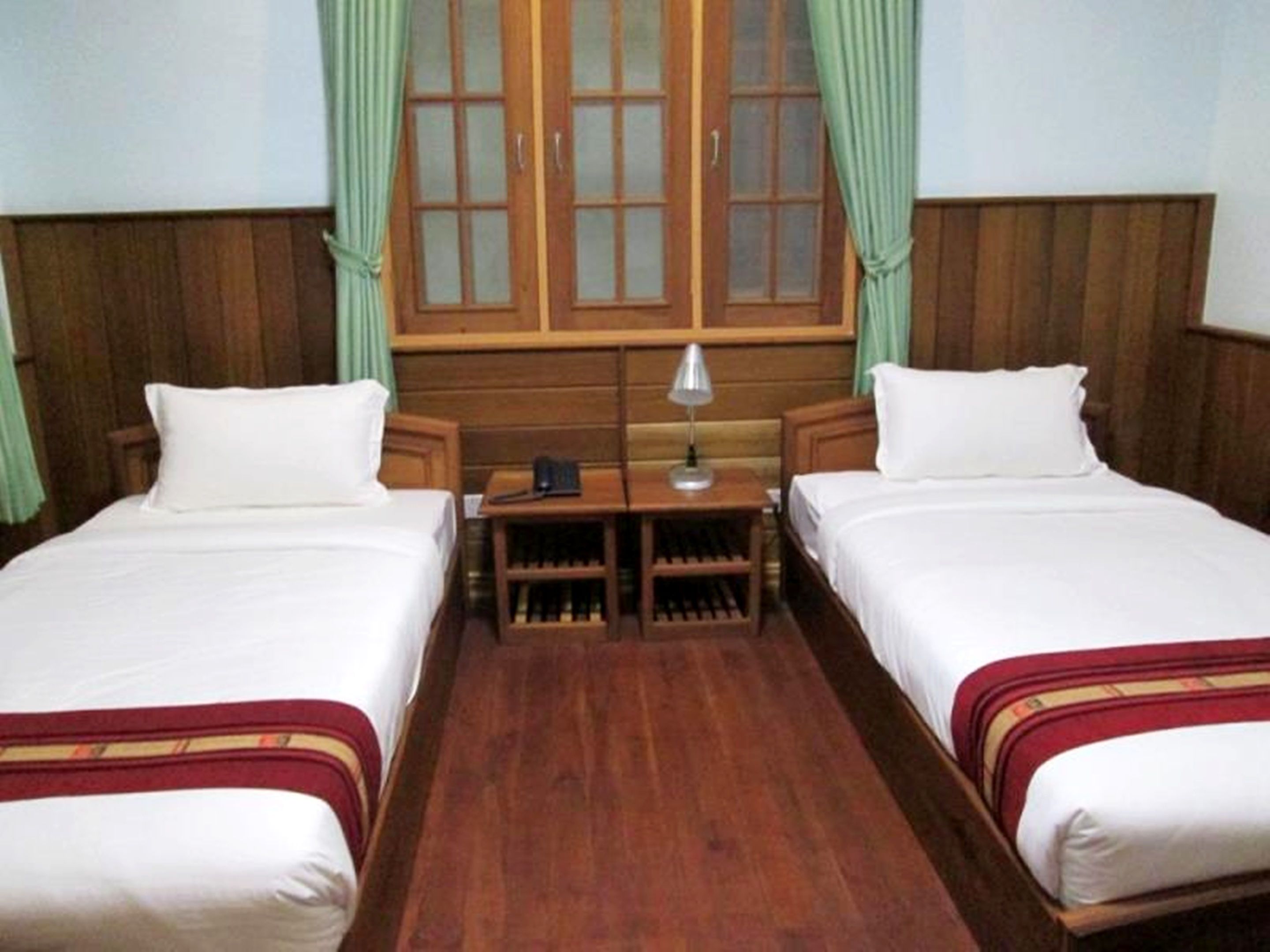 Una habitación con dos camas individuales, una mesita con lámpara y un ventanal con cortinas.