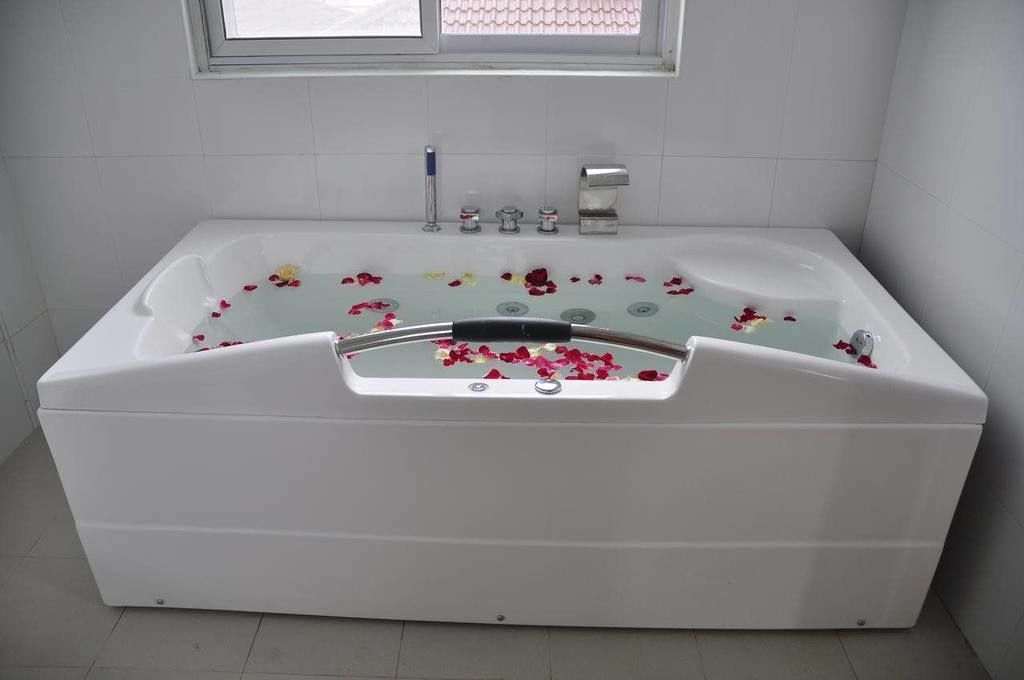 Una bañera blanca con pétalos de rosa flotando en el agua, una ventana arriba y una pared de azulejos blancos.