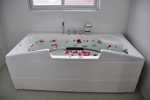 Una bañera blanca con pétalos de rosa flotando en el agua, una ventana arriba y una pared de azulejos blancos.