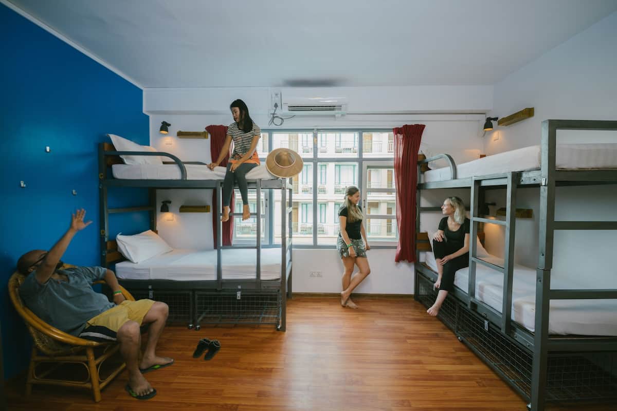 Une chambre de dortoir avec des lits superposés, une fenêtre avec des rideaux, un ventilateur sur pied et une personne assise sur une chaise.