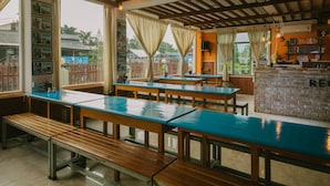 A restaurant with wooden tables and benches, large windows with curtains, and a brick counter area. - Ostello Bello Nyaung Shwe (Nyaungshwe)
